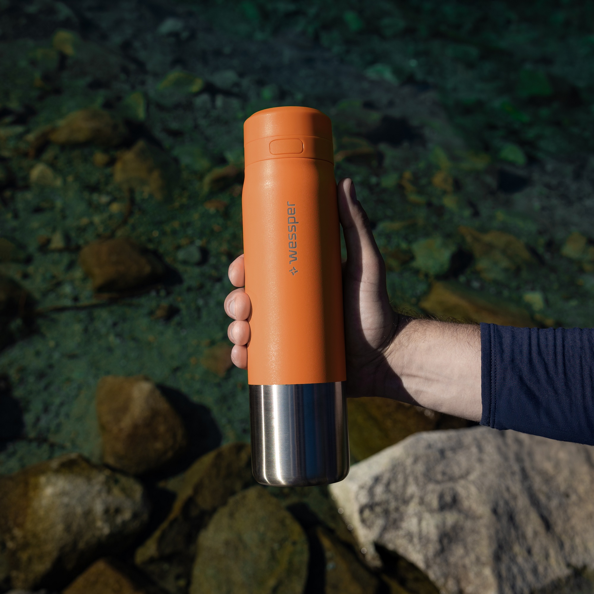 Prezentacja pomarańczowej butelki Wessper ze stali nierdzewnej na tle górskiego czystego strumienia